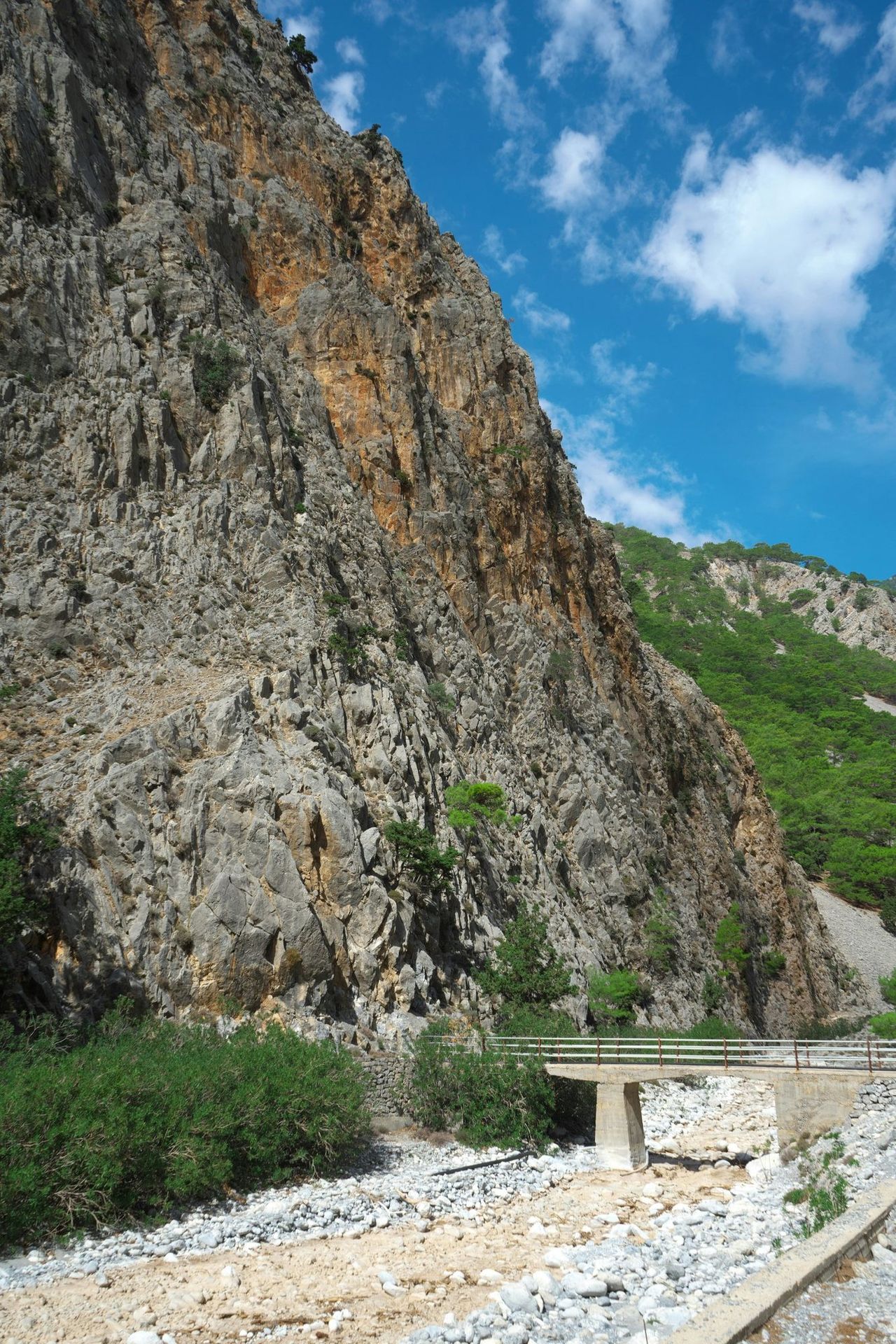 A small bridge over the dry riverbed under a high rock wall in the Samaria gorge