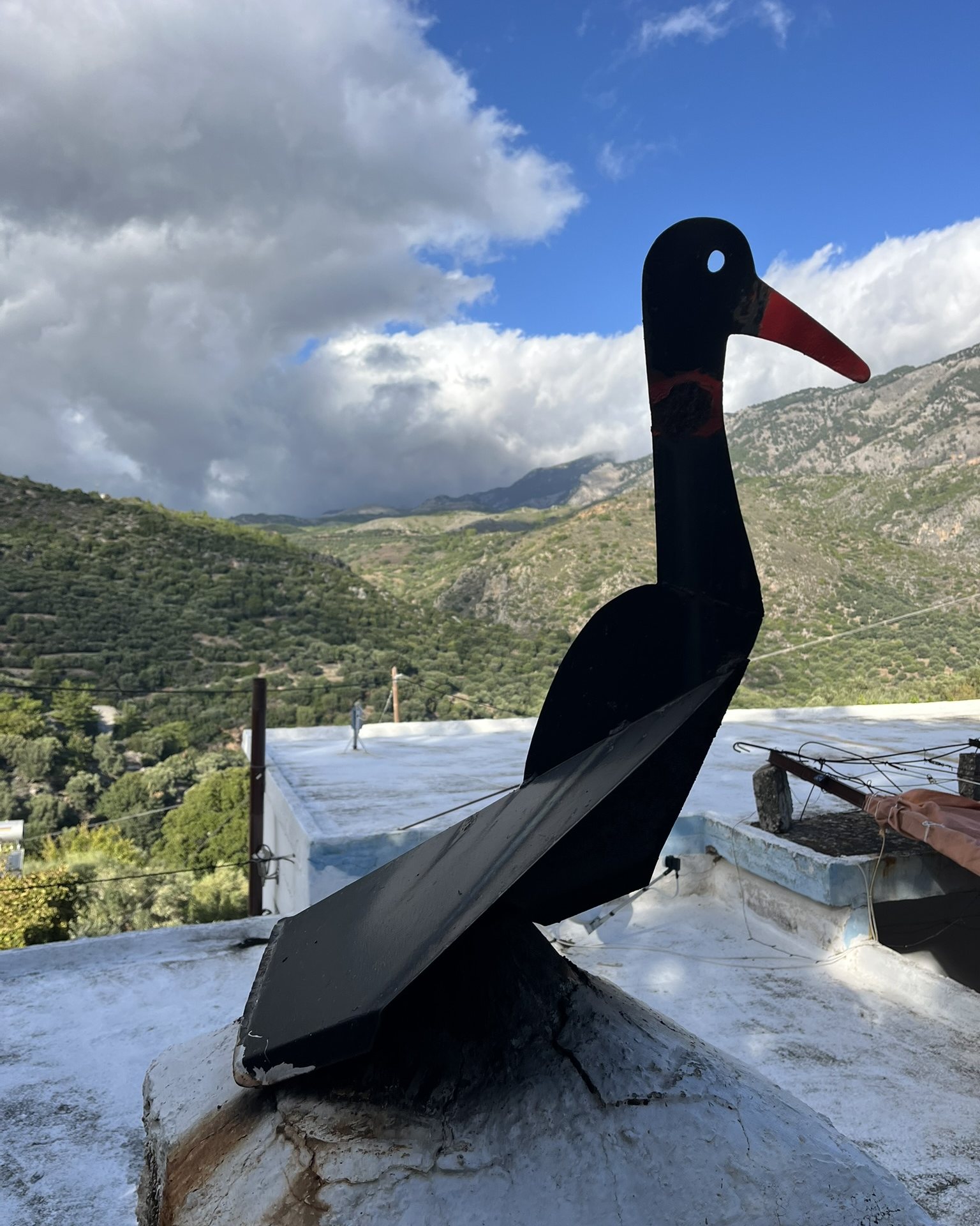 A black metal bird with a red beak on a white chimney in Moni, mountains and clouds behind