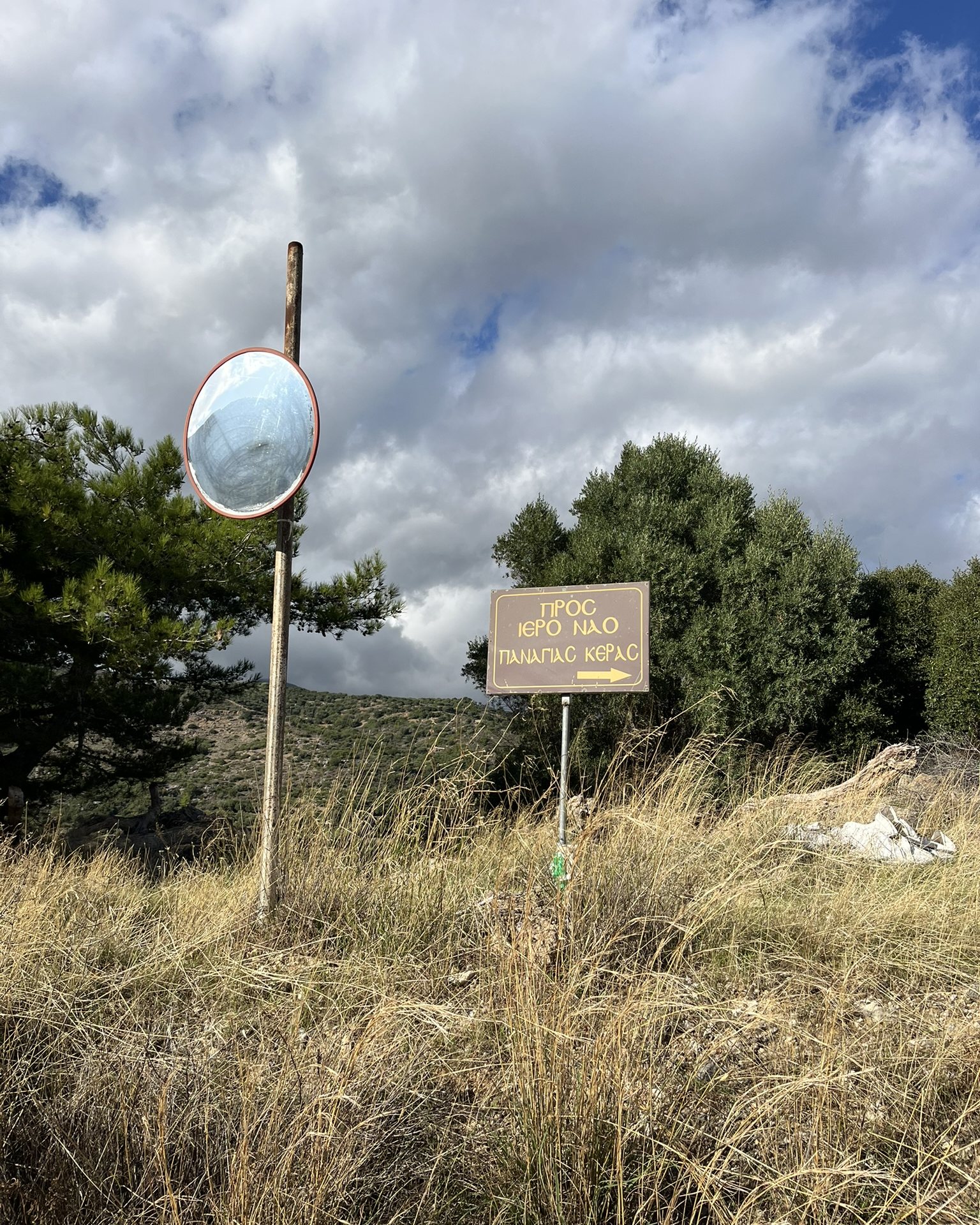 A brown sign in Greek pointing to the church of Panagia Kera, beside a road mirror and dry grass