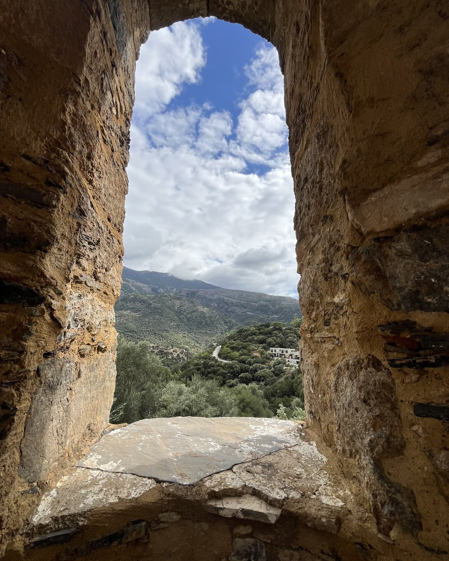A tall stone window in the bell tower framing the mountains, olive trees and a cloudy sky